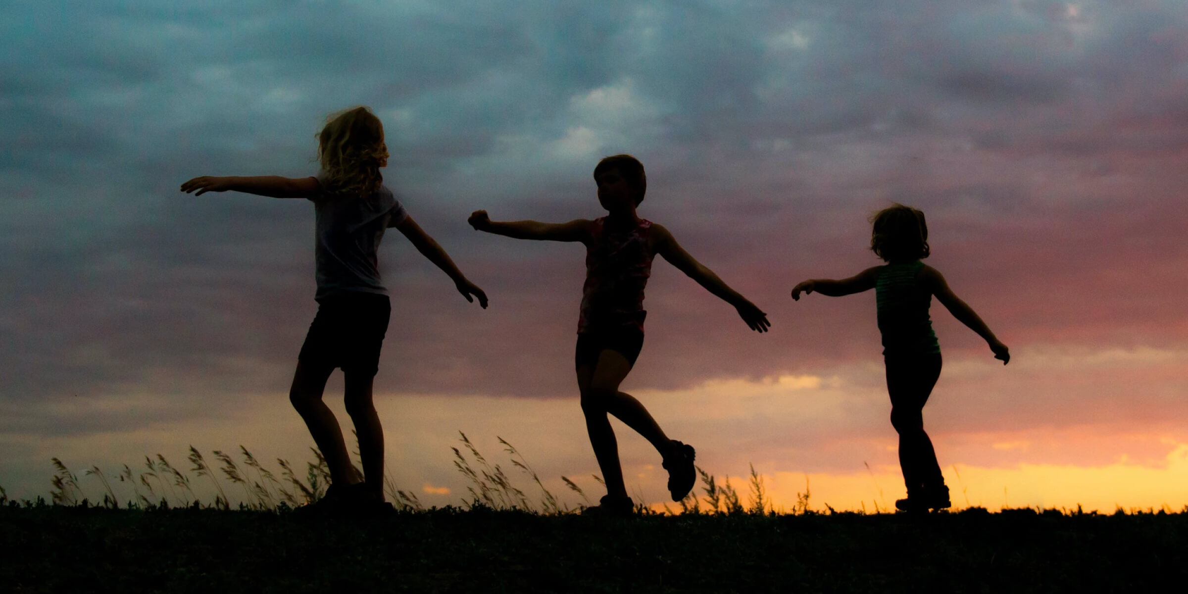 Silhouettes of three children playing outdoors at sunset under a cloudy sky